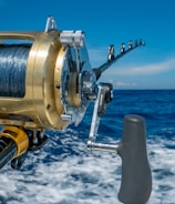 A close-up view of a fishing rod and reel on a boat, angling out over a deep blue ocean with a clear sky and a few clouds. The reel is shiny, metallic, and well-crafted, indicating it is likely for deep-sea fishing.