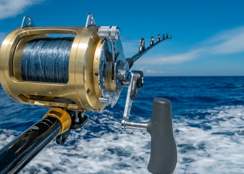 A close-up view of a fishing rod and reel on a boat, angling out over a deep blue ocean with a clear sky and a few clouds. The reel is shiny, metallic, and well-crafted, indicating it is likely for deep-sea fishing.