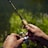 Hands holding a fishing rod with a wooden grip, situated near a body of water and surrounded by greenery, indicating an outdoor fishing scene.