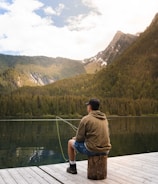 A person sits on a wooden dock by a serene mountain lake, casually fishing. They wear a hoodie, shorts, and a cap, and the rod extends into the water. The backdrop is a picturesque view of tree-covered mountains under a partly cloudy sky.