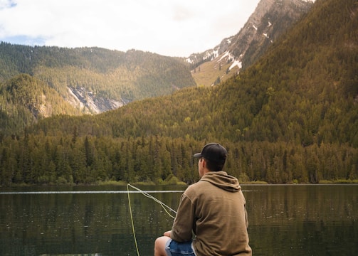 A person sits on a wooden dock by a serene mountain lake, casually fishing. They wear a hoodie, shorts, and a cap, and the rod extends into the water. The backdrop is a picturesque view of tree-covered mountains under a partly cloudy sky.