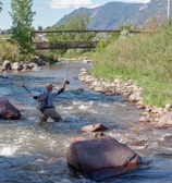 A person standing in a flowing stream, holding a fishing rod in one hand and a net in the other. The surrounding landscape includes lush greenery and rocky banks, with a metal bridge and distant mountains visible in the background under a clear blue sky.