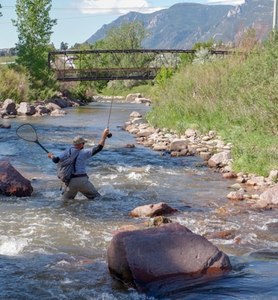 A person standing in a flowing stream, holding a fishing rod in one hand and a net in the other. The surrounding landscape includes lush greenery and rocky banks, with a metal bridge and distant mountains visible in the background under a clear blue sky.