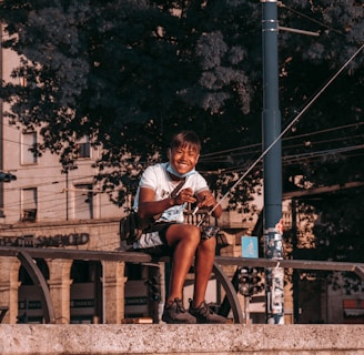 A person sitting on a stone ledge, smiling while holding a fishing rod. The background consists of large, leafy trees and a section of an urban environment, including buildings and street elements such as a utility pole.