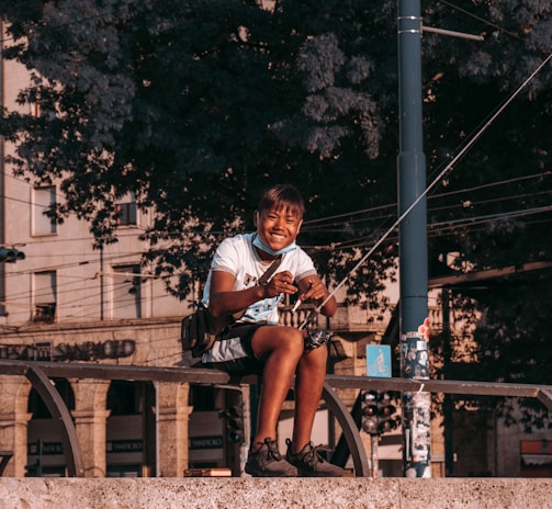 A person sitting on a stone ledge, smiling while holding a fishing rod. The background consists of large, leafy trees and a section of an urban environment, including buildings and street elements such as a utility pole.