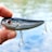A hand holding a shiny fishing lure with red eyes and two treble hooks. The background shows a blurred view of water, suggesting an outdoor setting.