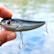 A hand holding a shiny fishing lure with red eyes and two treble hooks. The background shows a blurred view of water, suggesting an outdoor setting.