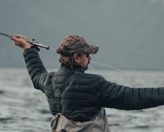 A person wearing a camouflage cap and a black puffy jacket is holding a fishing rod, actively casting a line into a large body of water. The background is composed of blurred mountains and a cloudy sky, conveying an outdoor, nature setting.