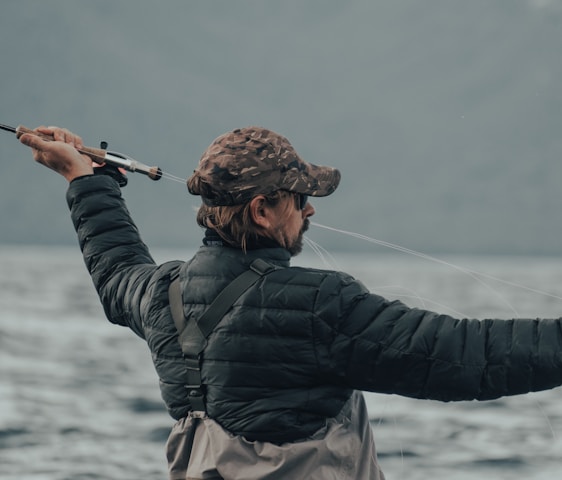 A person wearing a camouflage cap and a black puffy jacket is holding a fishing rod, actively casting a line into a large body of water. The background is composed of blurred mountains and a cloudy sky, conveying an outdoor, nature setting.