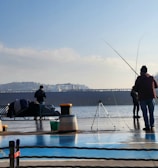 Several people are standing at a waterfront with fishing rods, engaging in fishing activities. A large cargo ship is visible in the background, sailing across the water. The scene is calm with clear skies, and there are various fishing gear items and a bench nearby.