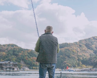 A person is fishing by a waterfront, sitting on a stool with scenic hills in the background. The setup includes various containers and equipment nearby, indicating preparation for a fishing activity.