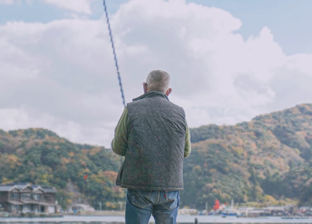 A person is fishing by a waterfront, sitting on a stool with scenic hills in the background. The setup includes various containers and equipment nearby, indicating preparation for a fishing activity.