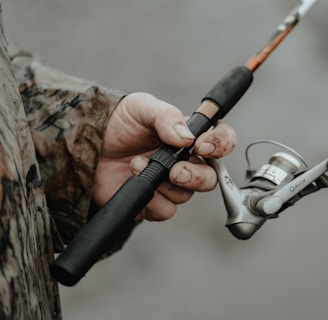 A person wearing a camouflage jacket is holding a fishing rod. The fishing reel is prominently featured, with the hand showing some dirt and wear, suggesting an outdoor activity.