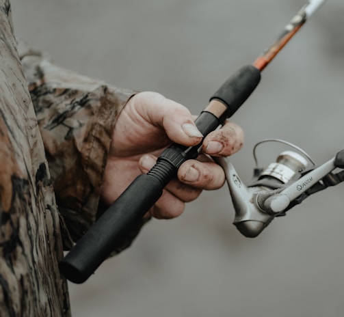 A person wearing a camouflage jacket is holding a fishing rod. The fishing reel is prominently featured, with the hand showing some dirt and wear, suggesting an outdoor activity.