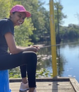 A person is sitting on a blue stool near a body of water, holding a fishing rod. The individual is wearing a pink cap, black t-shirt, and leggings, smiling while facing the water. The surroundings are lush with greenery, and there is a tranquil river reflecting the blue sky and trees.