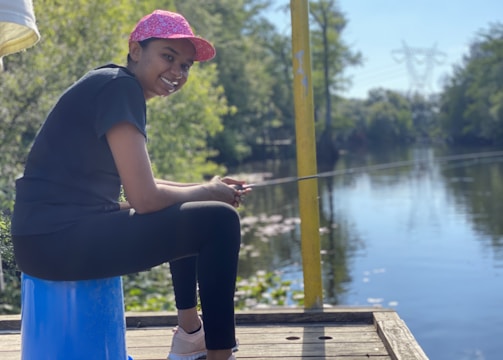 A person is sitting on a blue stool near a body of water, holding a fishing rod. The individual is wearing a pink cap, black t-shirt, and leggings, smiling while facing the water. The surroundings are lush with greenery, and there is a tranquil river reflecting the blue sky and trees.