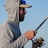 A person wearing a light gray hoodie and a cap is holding a fishing rod. The sun reflects on their sunglasses, and they appear to be focused on fishing. The background shows a clear blue sky.
