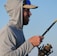 A person wearing a light gray hoodie and a cap is holding a fishing rod. The sun reflects on their sunglasses, and they appear to be focused on fishing. The background shows a clear blue sky.