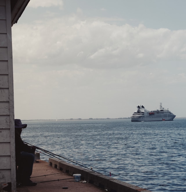 A person sits on a dock, fishing with a rod while a large cruise ship sails in the background over the calm ocean waters. The sky is partly cloudy, and the scene conveys a sense of tranquility and leisure.