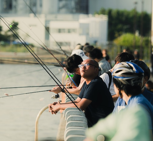 Several people are lined up along a railing by a body of water, engaged in fishing. Each person holds a fishing rod, with the lines cast into the water. The background shows an industrial or urban setting with buildings and greenery. The scene captures both leisure and concentration.