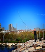 A person is fishing on a rocky embankment near a body of water, with a backdrop of a cityscape featuring modern buildings. The sky is clear and blue, and a group of people with bicycles is nearby, suggesting a leisurely, outdoor setting.
