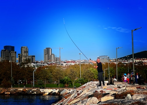 A person is fishing on a rocky embankment near a body of water, with a backdrop of a cityscape featuring modern buildings. The sky is clear and blue, and a group of people with bicycles is nearby, suggesting a leisurely, outdoor setting.