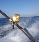 A fishing rod with a gold reel is mounted on a holder as a boat speeds over the ocean, causing splashes of water and creating a sense of motion. The sky above is clear and blue, and a rainbow is faintly visible in the spray.