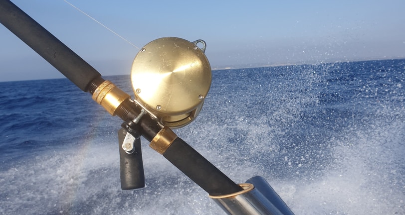 A fishing rod with a gold reel is mounted on a holder as a boat speeds over the ocean, causing splashes of water and creating a sense of motion. The sky above is clear and blue, and a rainbow is faintly visible in the spray.